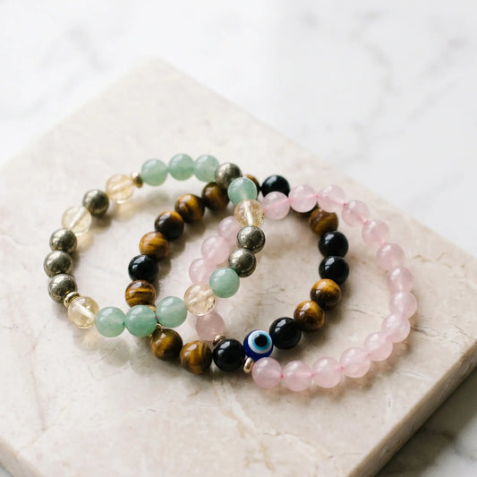 "Three premium 8mm natural gemstone bracelets—Dhan Yog, Triple Eye Protection, and Rose Quartz—elegantly resting together on a light marble slab in soft natural daylight."