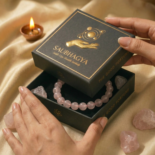 Hands opening a premium black Saubhagya gift box revealing a natural 8mm Rose Quartz gemstone bracelet. The luxury unboxing setting includes raw rose quartz crystals and a warm lit diya.