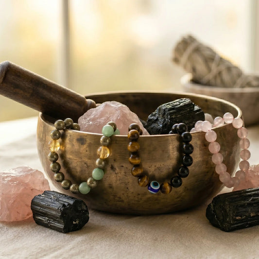 A still-life photograph featuring a weathered bronze singing bowl on a textured linen cloth, filled with raw rose quartz and black tourmaline crystals. Several beaded bracelets made of pyrite, tiger's eye (with an evil eye), and rose quartz are draped over the rim. A dark wooden striker rests on the edge. More raw crystals and a bundle of white sage are in the softly blurred, sunlit background. The lighting is warm and natural, creating a meditative feel.